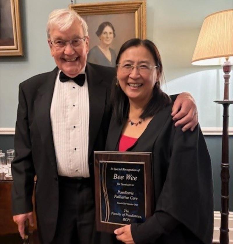 A woman in glasses and black gown holds an award, next to a man in a tuxedo 