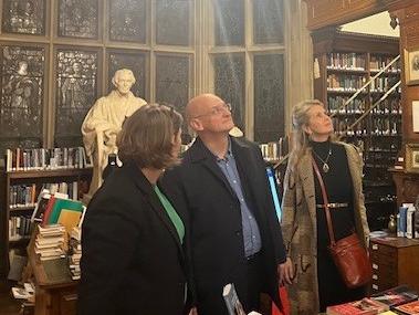 A man looks up in a college library. There is a statue behind him, and two women stand, one on each side of him, also looking up.
