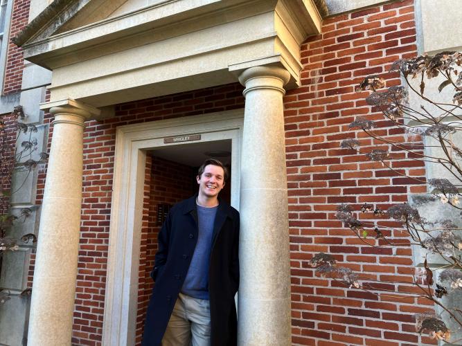 A young man leans against the column of a red-brick building