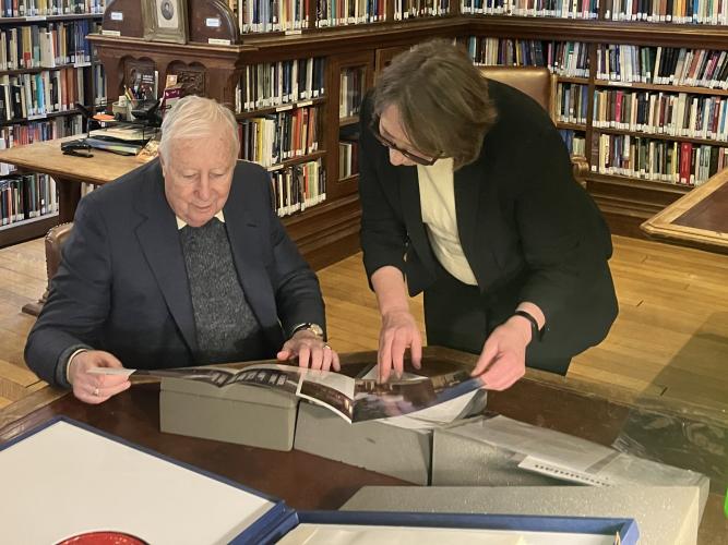 An elderly man sitting down inspecting materials in a library with a woman standing over him