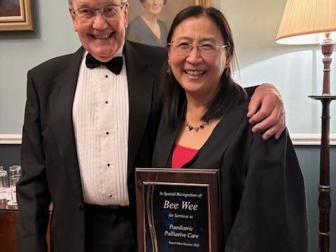 A woman in glasses and black gown holds an award, next to a man in a tuxedo 