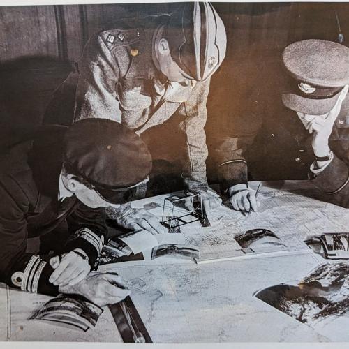 A black and white photograph of three men in military uniforms leaning over photographs and maps 