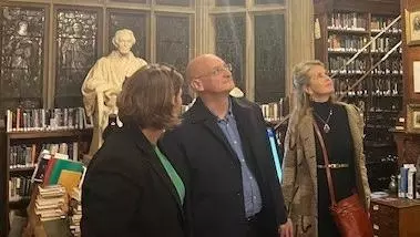 A man looks up in a college library. There is a statue behind him, and two women stand, one on each side of him, also looking up.