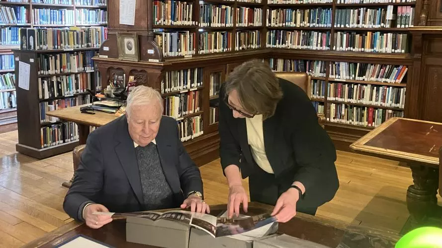 An elderly man sitting down inspecting materials in a library with a woman standing over him