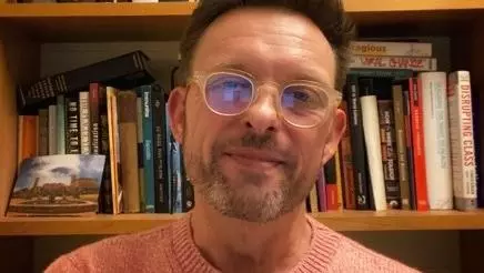 Portrait of Professor Matthew Weait smiling and facing directly ahead. He is wearing a light orange jumper and glasses, and is sitting in front of a bookcase.