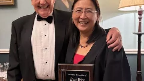 A woman with glasses and gown, next to a man in a tuxedo with his arm around her, holds an award