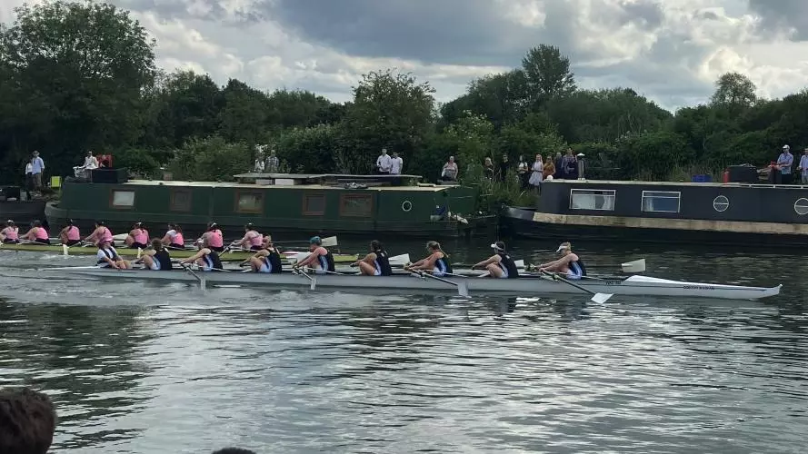 Two rowing boats on a river with canal boats and supporters in the background
