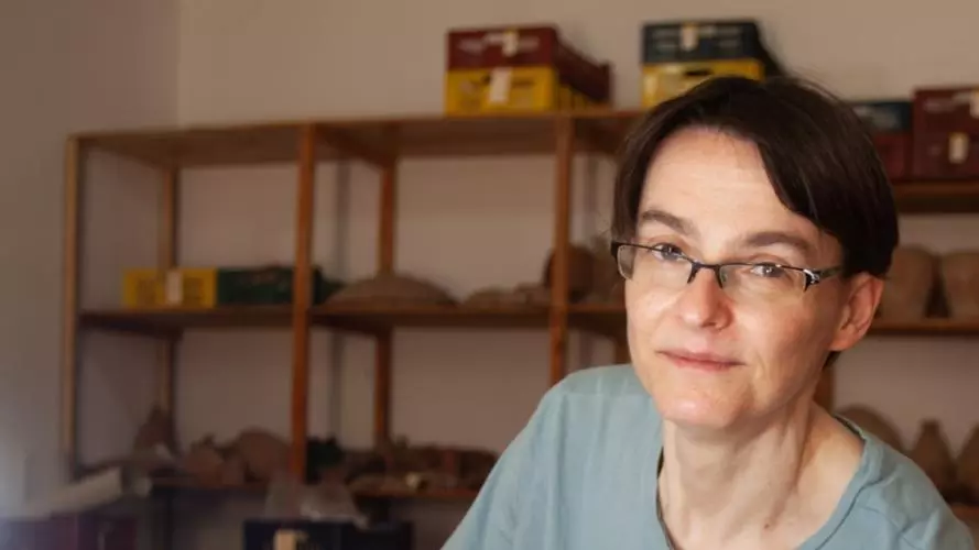 Photo portrait of Hana Navratilova, waring a t-shirt, with archaeological finds on shelves behind