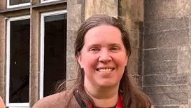 A woman wearing a brown cardigan standing in front of a building with windows.