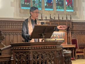Prof Santanu Das speaking at the lectern of the college chapel with one hand outstretched