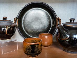 Two teapots and a plate in a dark glaze with two cups in the foreground