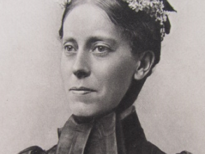 Picture of a women (headshot) with smiling with a headress with flowers and dressed in black victorian clothes