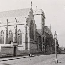 A black and white photograph of a street scene with the College buildings to the left