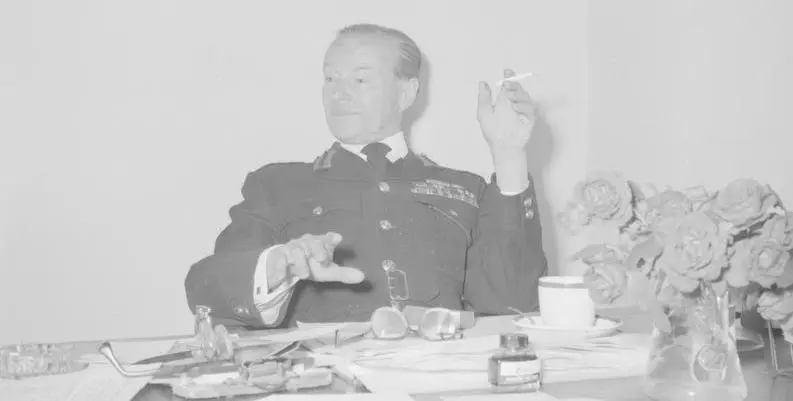 A man sitting in uniform behind a desk, leaning back and smoking a cigarette.  On the desk there are papers and flowers.  