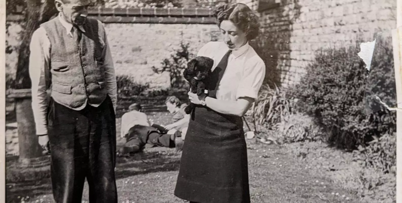 Two figures standing in a walled garden.  The female figure is holding a black puppy.  The photo was taken in 1945.