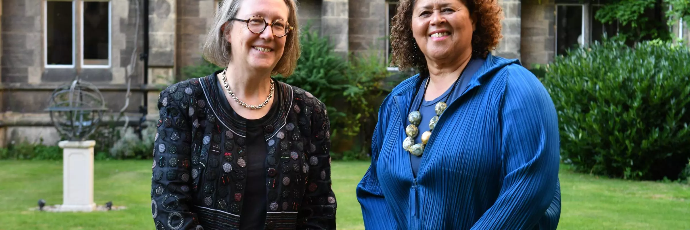 The Principal and Anna Deavere Smith smiling together in the Tate Quad. In the background, stonework, grass and green foliage